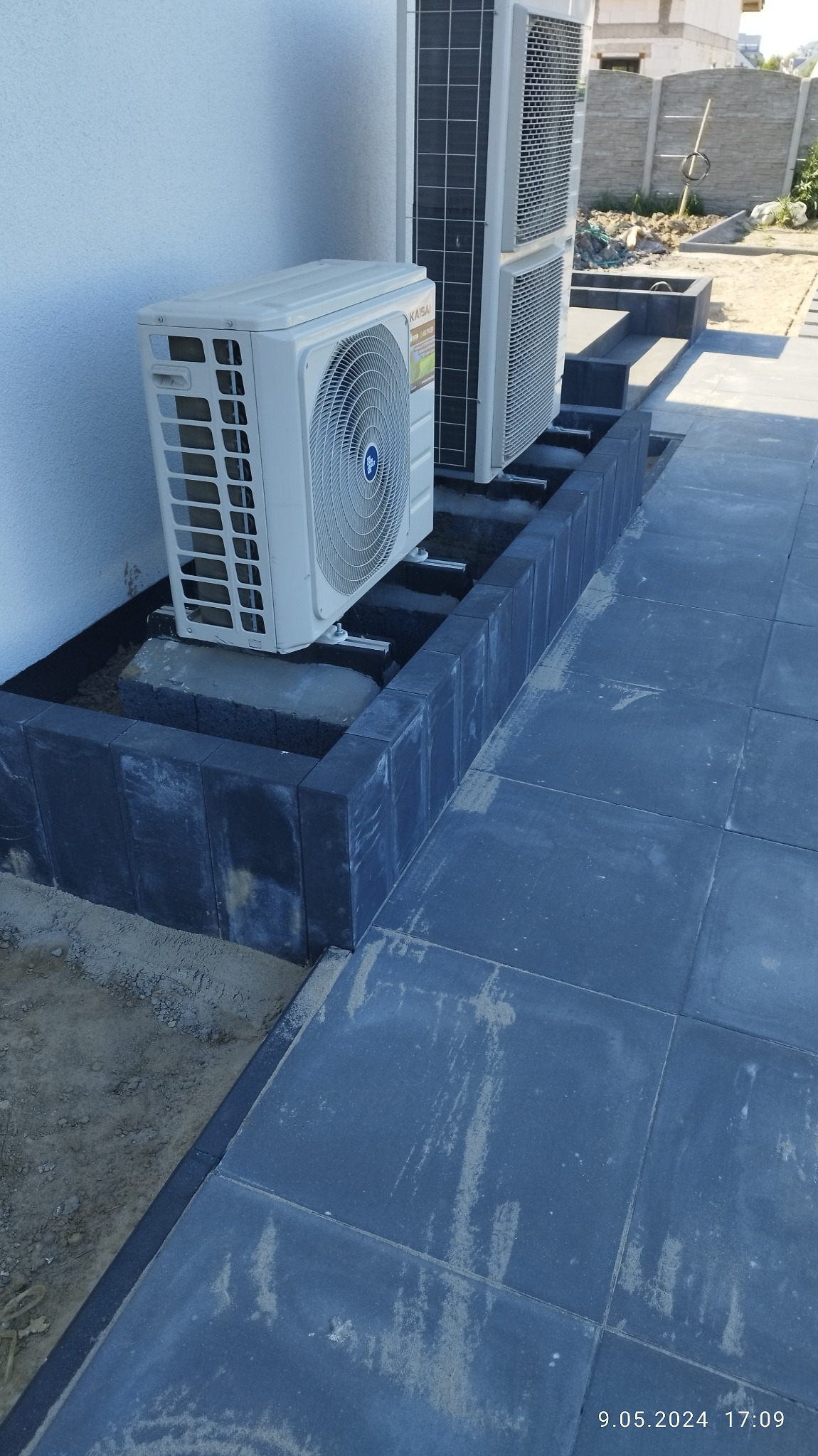 Szare płyty chodnikowe ułożone przy ścianie budynku, z obrzeżem z ciemnych kostek, tworzącym podstawę pod klimatyzatory.