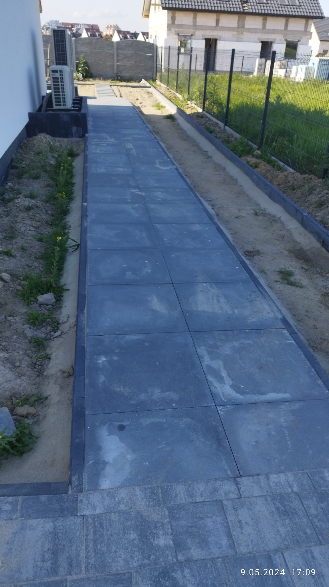 Długa, prosta ścieżka z ciemnoszarych, kwadratowych płyt brukowych prowadząca wzdłuż białej ściany budynku i ogrodzenia z siatki, z widocznym klimatyzatorem na ścianie i fragmentem trawnika.