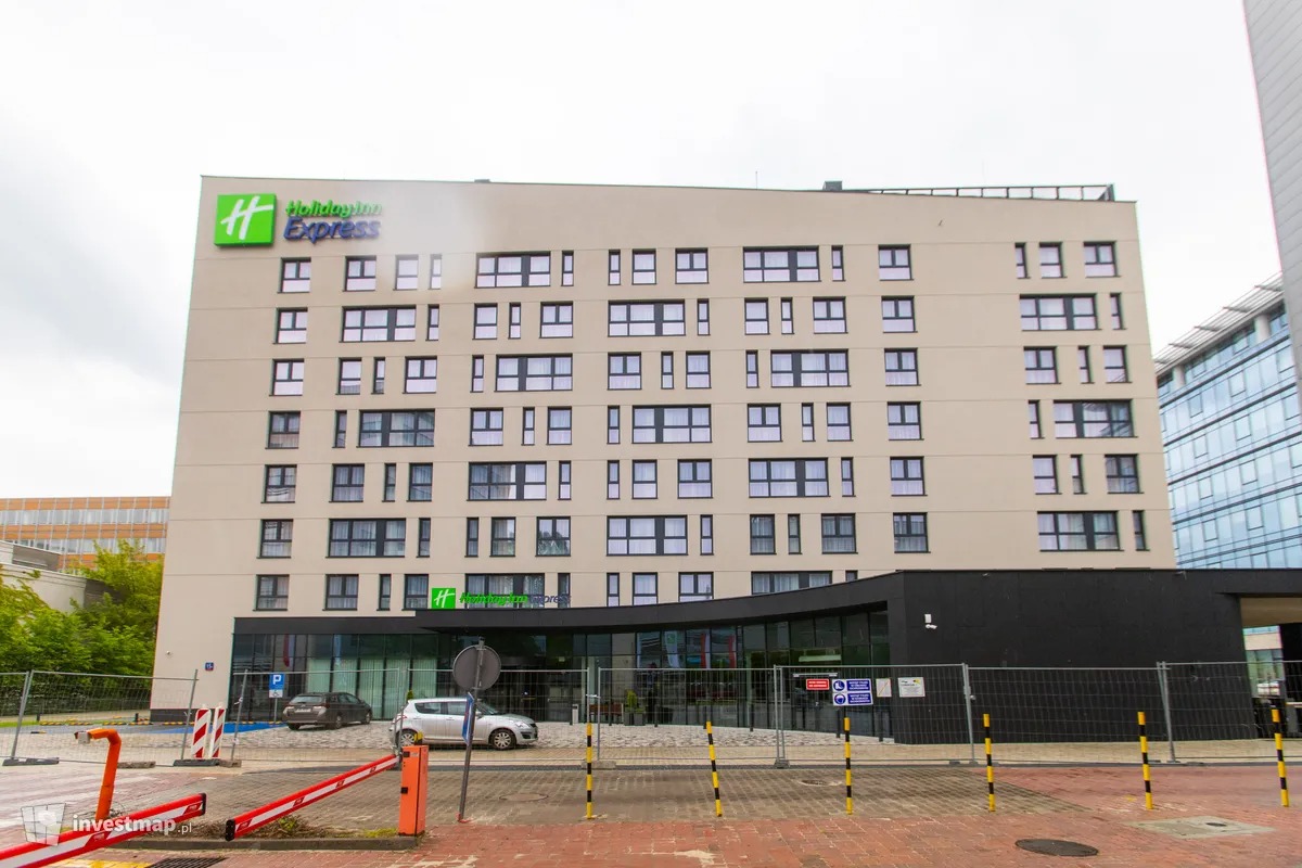 Wielopiętrowy budynek hotelowy z logo Holiday Inn Express, widoczny z poziomu ulicy, z zaparkowanymi samochodami i barierkami ochronnymi.