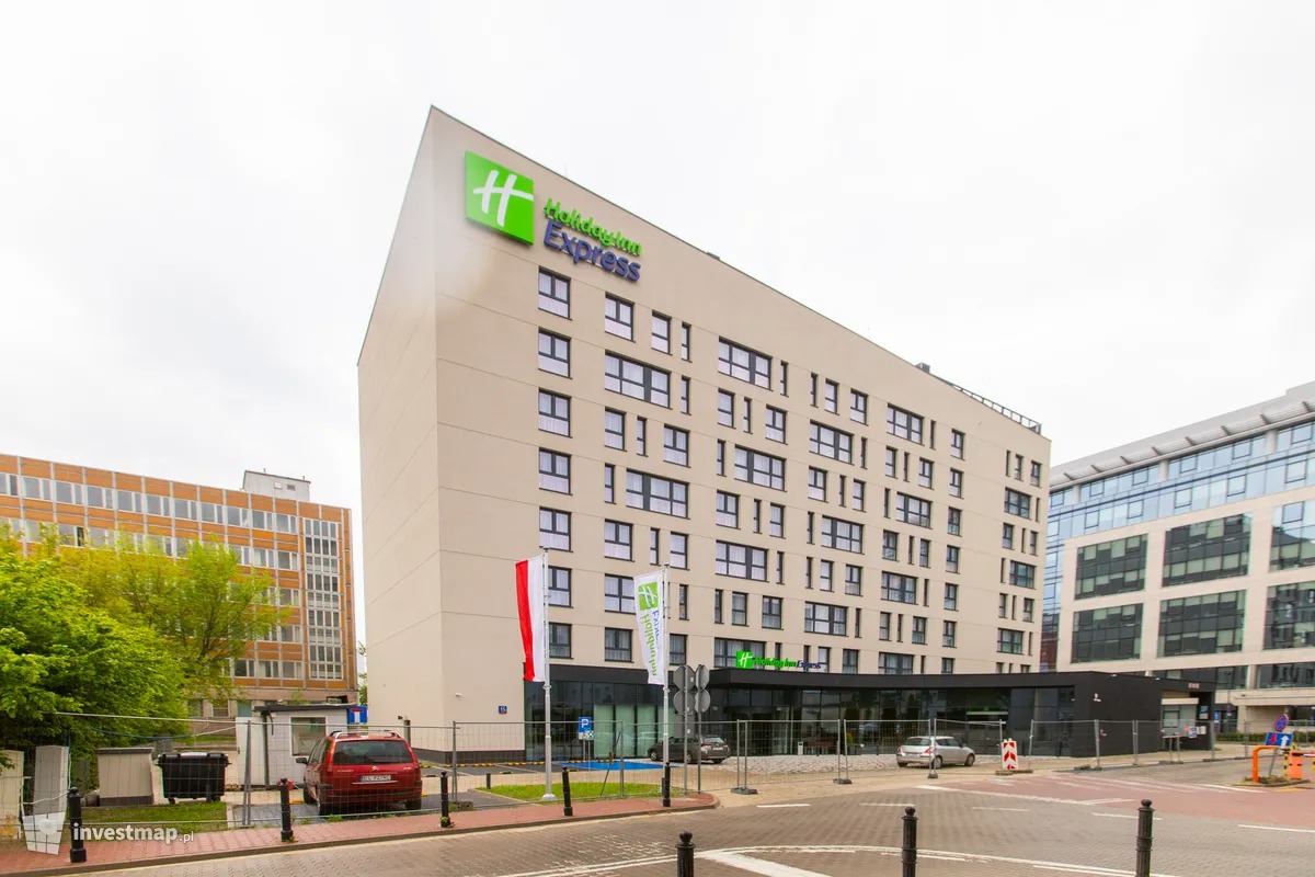 Nowoczesny, wielopiętrowy budynek hotelowy z płaskim dachem i zielonym logo sieci, widoczny z poziomu ulicy, otoczony innymi budynkami i drzewami.