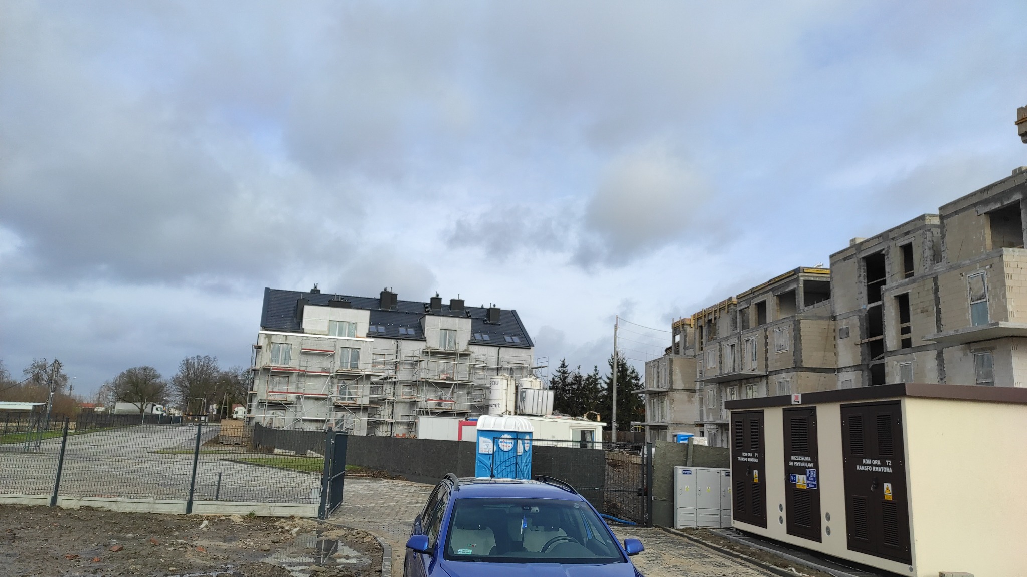 Wielorodzinny budynek w trakcie budowy, widoczne rusztowania, stan surowy zamknięty z oknami, na pierwszym planie niebieski samochód i toaleta przenośna.