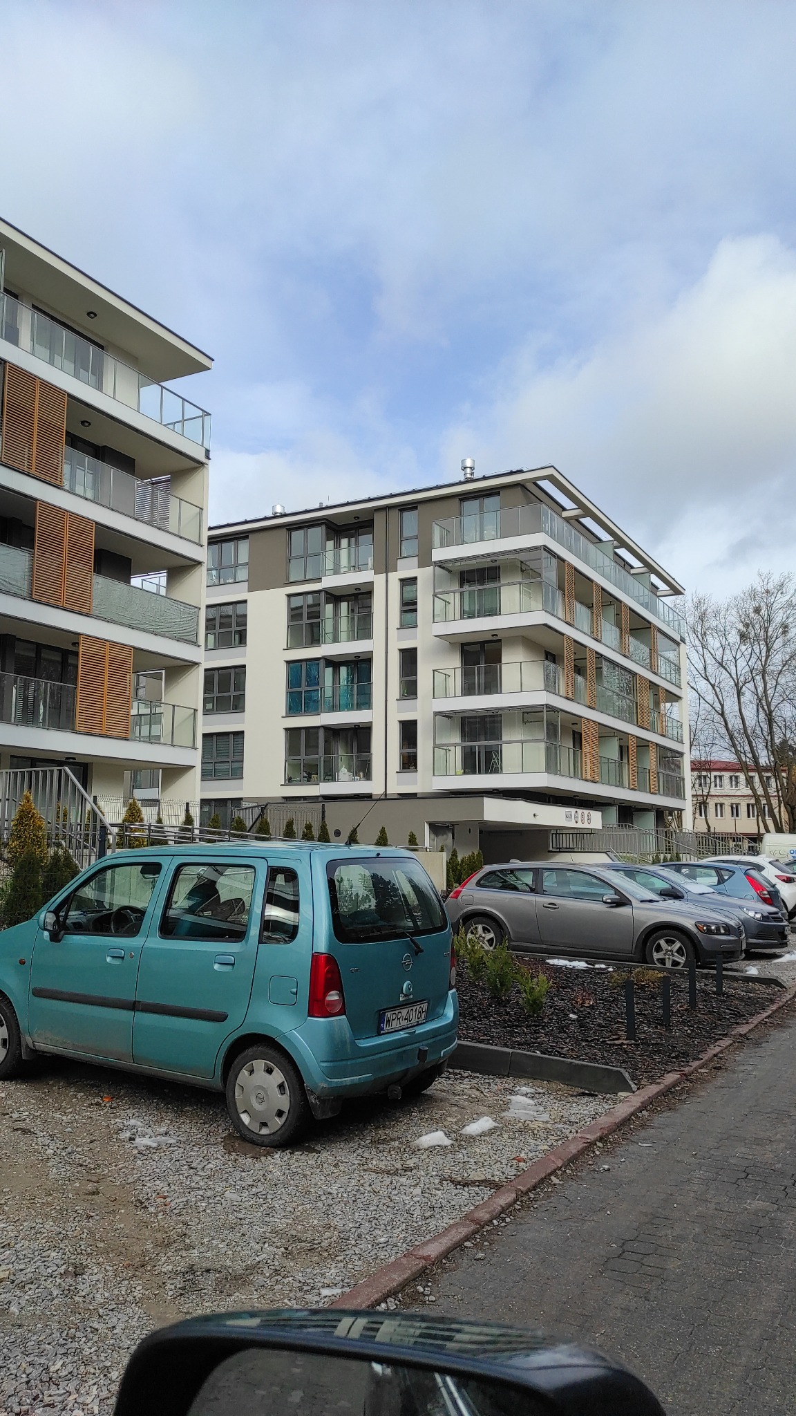 Nowoczesny budynek wielorodzinny z balkonami i szklanymi balustradami, widok z parkingu z zaparkowanymi samochodami w pochmurny dzień.