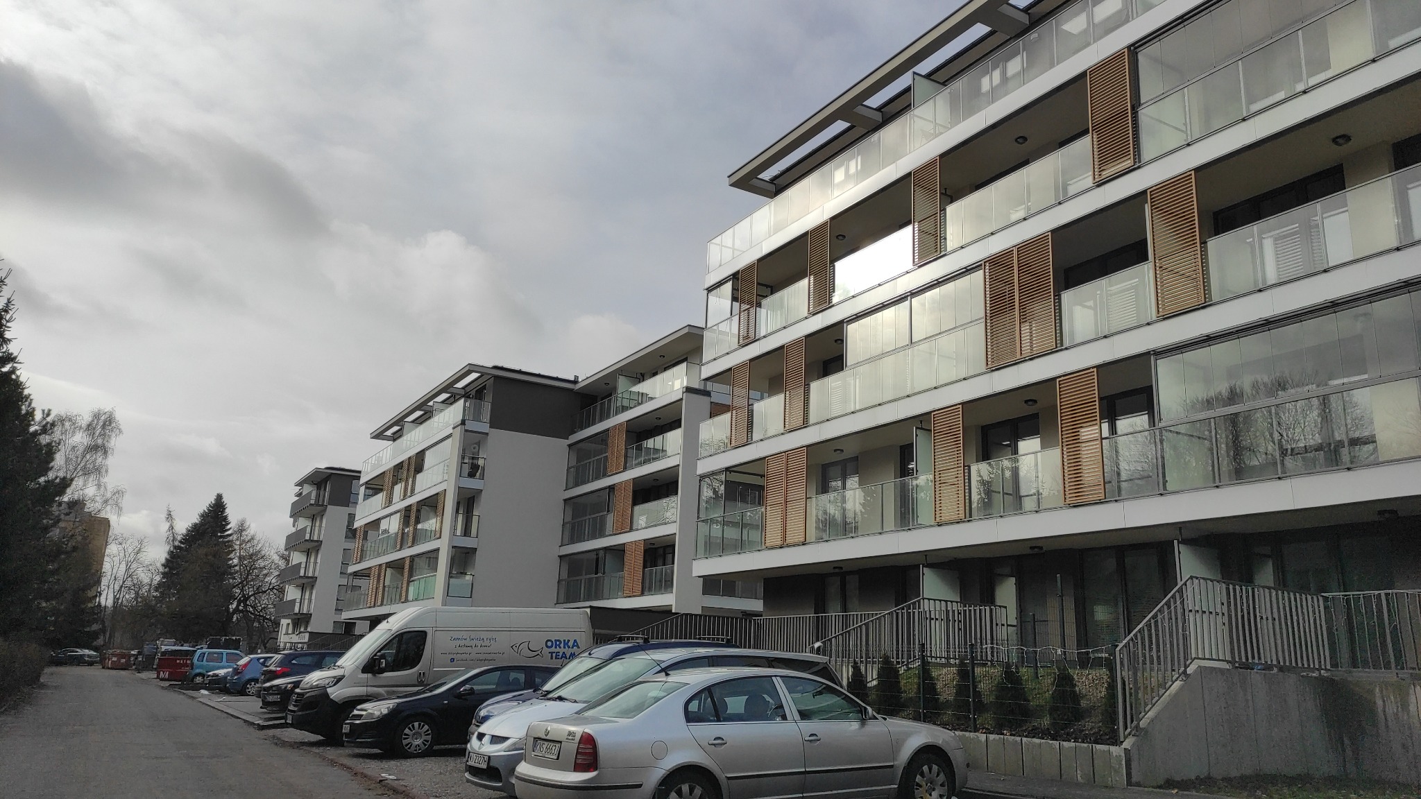 Nowoczesny budynek żelbetowy z balkonami z drewnianymi żaluzjami, widok z parkingu z zaparkowanymi samochodami i furgonetką firmy budowlanej Orka Team.