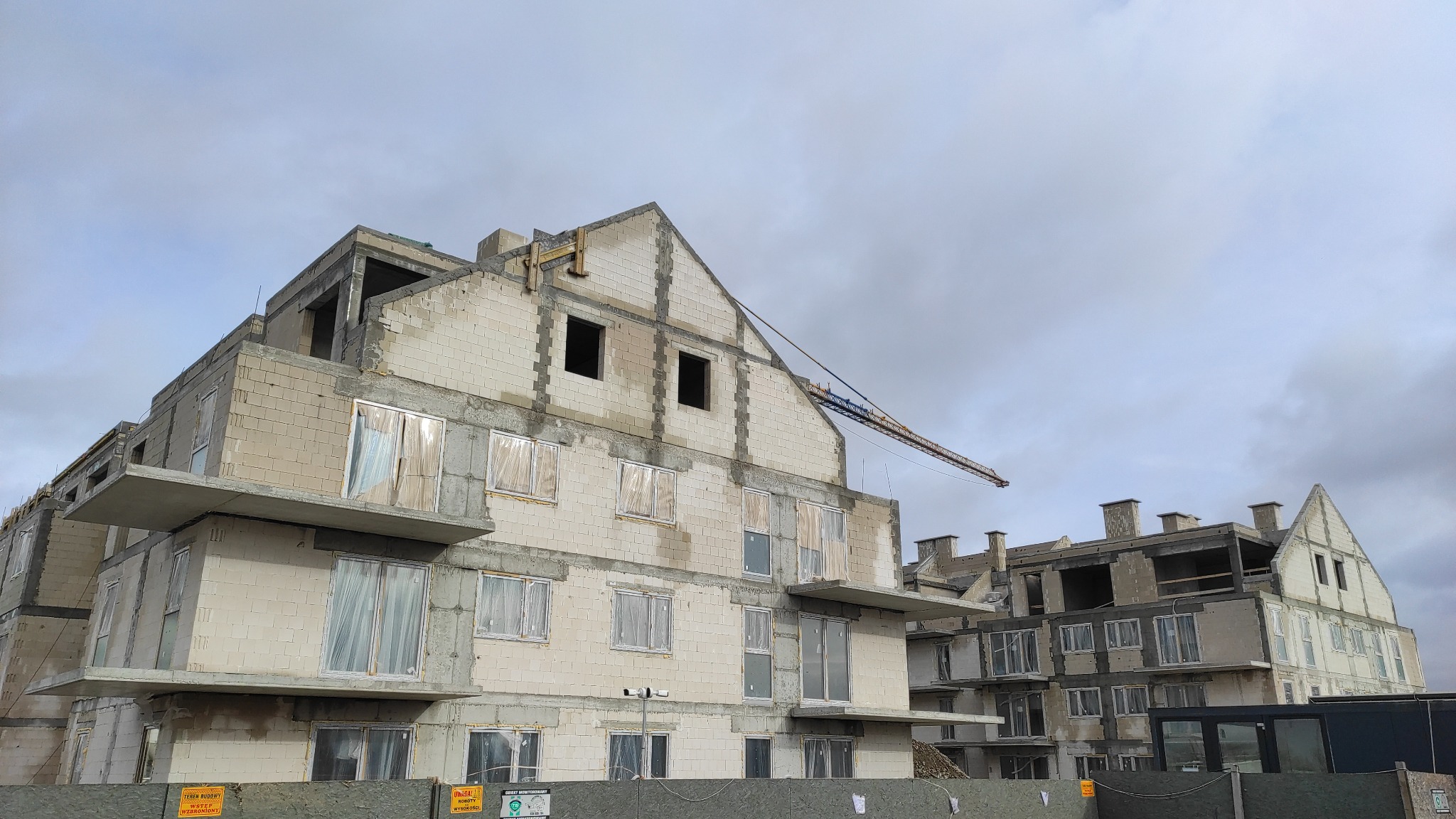 Surowy stan wielopiętrowego budynku z betonu komórkowego, z widocznym żurawiem budowlanym i zabezpieczonymi folią oknami, na tle pochmurnego nieba.