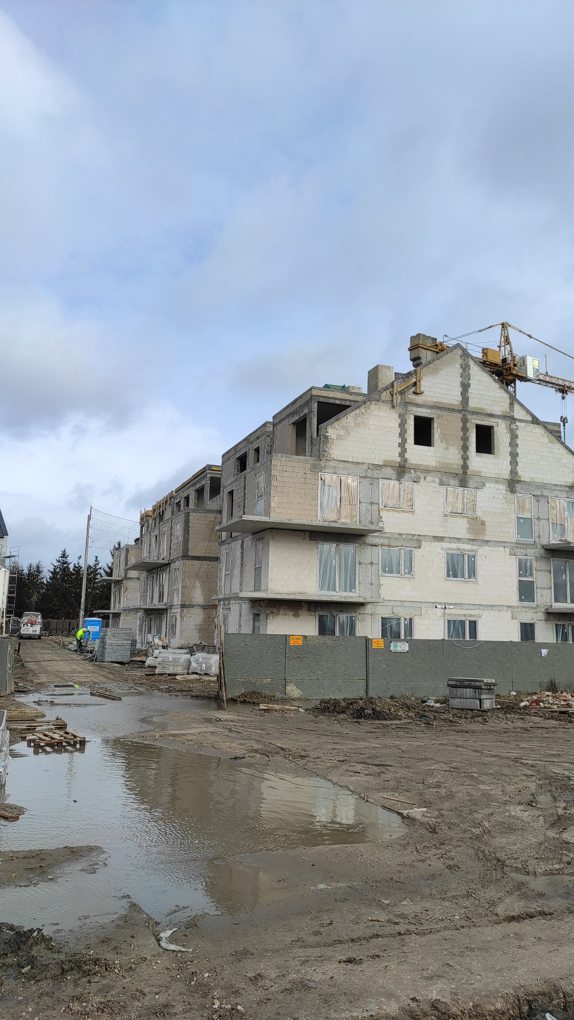 Wznoszenie budynku wielorodzinnego z betonu komórkowego w Warszawie, widoczny żuraw wieżowy i materiały budowlane składowane na terenie budowy, po deszczu.