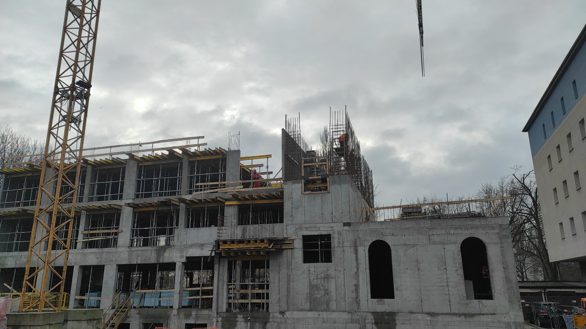 Wznoszenie wielopiętrowego budynku w konstrukcji żelbetowej z widocznym żurawiem, szalunkami i zbrojeniami na tle pochmurnego nieba.