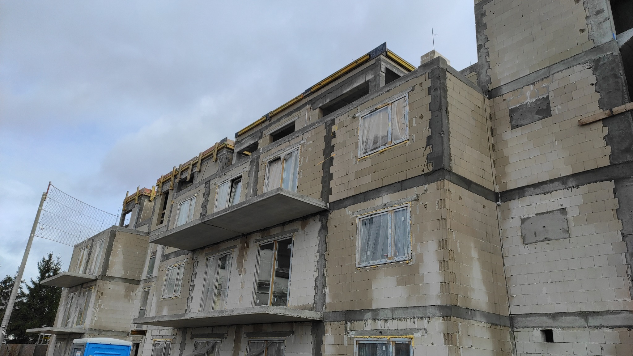 Wznoszenie surowej bryły budynku wielorodzinnego z betonu komórkowego, widoczne balkony i szalunki stropowe, okna zabezpieczone folią.