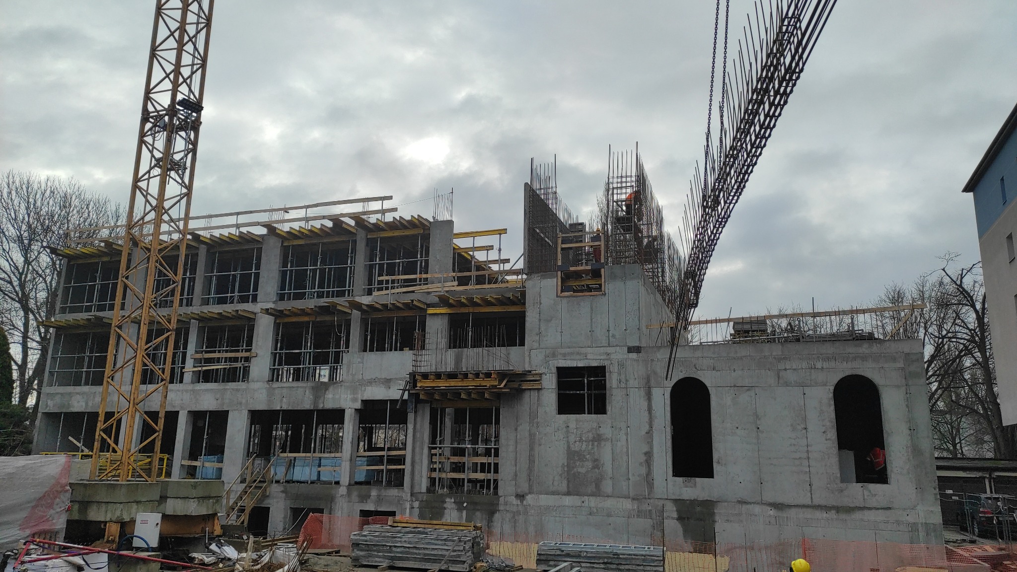 Wznoszenie wielopiętrowego budynku żelbetowego: szkielet konstrukcji z widocznym zbrojeniem, deskowaniem i dźwigiem w pochmurny dzień.