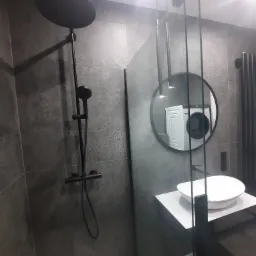 Nowoczesna łazienka z szarą, teksturowaną ścianą, czarną armaturą prysznicową typu deszczownica, szklaną kabiną prysznicową, okrągłym lustrem odbijającym pralkę oraz białą umywalką nablatową.