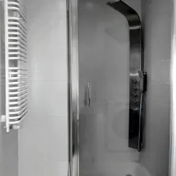 Nowoczesna kabina prysznicowa z panelem natryskowym w minimalistycznej łazience z białymi płytkami i grzejnikiem drabinkowym.