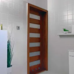 Nowoczesna łazienka z białymi kwadratowymi płytkami, drewnianą ramą drzwiową z matowymi szybami, minimalistyczną umywalką i dekoracyjnymi roślinami w doniczkach.