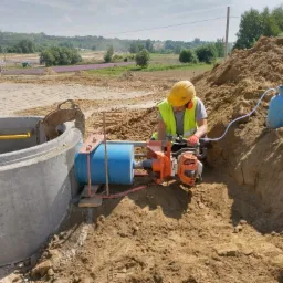 Pracownik w kasku i kamizelce odblaskowej wycina otwór w betonowym kręgu studni za pomocą wiertnicy diamentowej chłodzonej wodą, w tle widoczne pole i zabudowania.