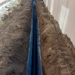 Długi, wąski wykop z czarną rurą drenażową ułożoną na geowłókninie i zasypaną kamieniami, widoczny fragment pracownika w odblaskowej kamizelce przy betonowym ogrodzeniu.