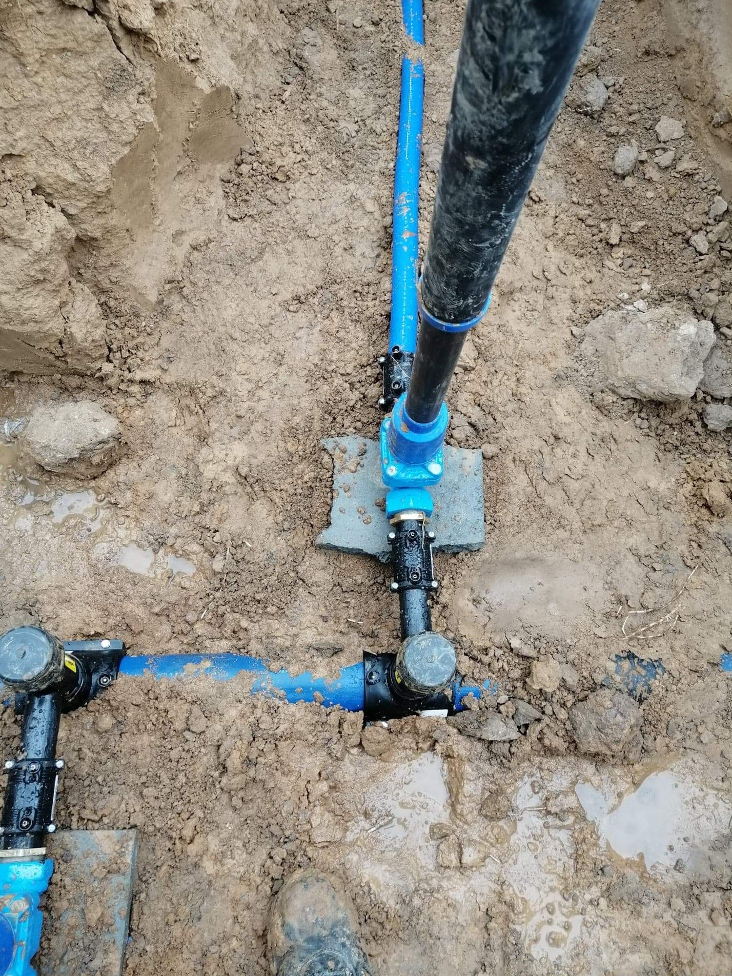 Niebieskie i czarne rury instalacyjne w wykopie z mokrą, gliniastą ziemią, widoczne połączenia i elementy złączek.