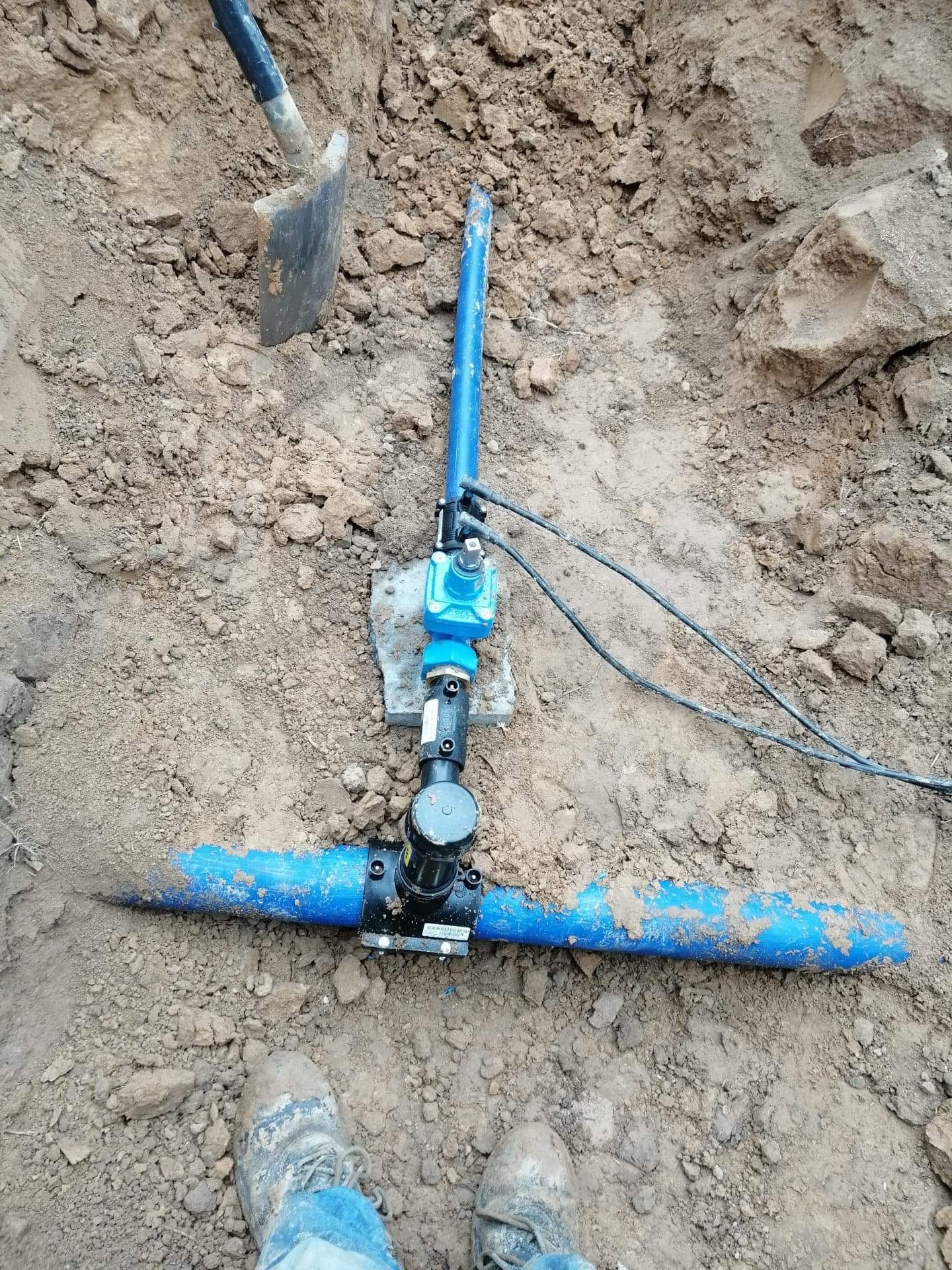 Fragment wykopu z ułożonym fragmentem instalacji wodnej z niebieskich rur z czarnymi złączkami, widoczna łopata i buty w ziemi.