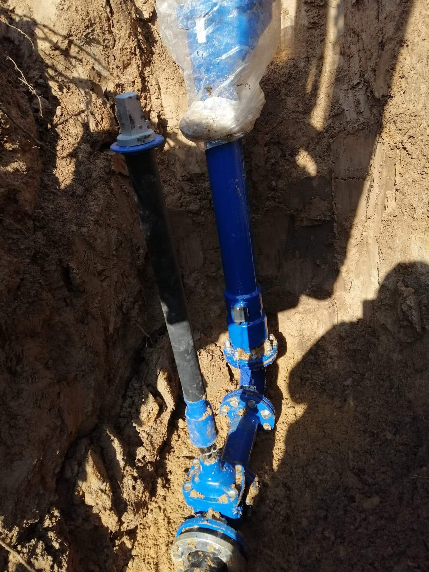 Niebieskie i czarne rury wodociągowe w wykopie ziemnym, z plastikową folią ochronną na jednym z przyłączy.