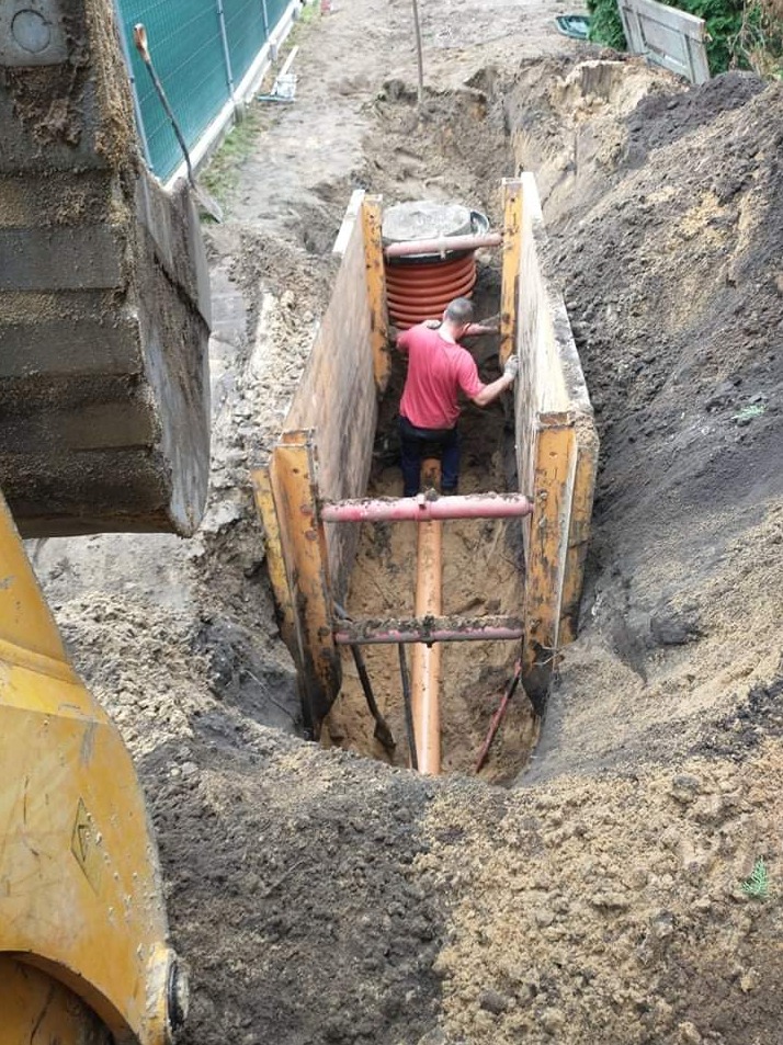 Pracownik w wykopie zabezpieczonym szalunkiem, przygotowuje się do montażu pomarańczowej rury kanalizacyjnej, widoczna koparka z boku.