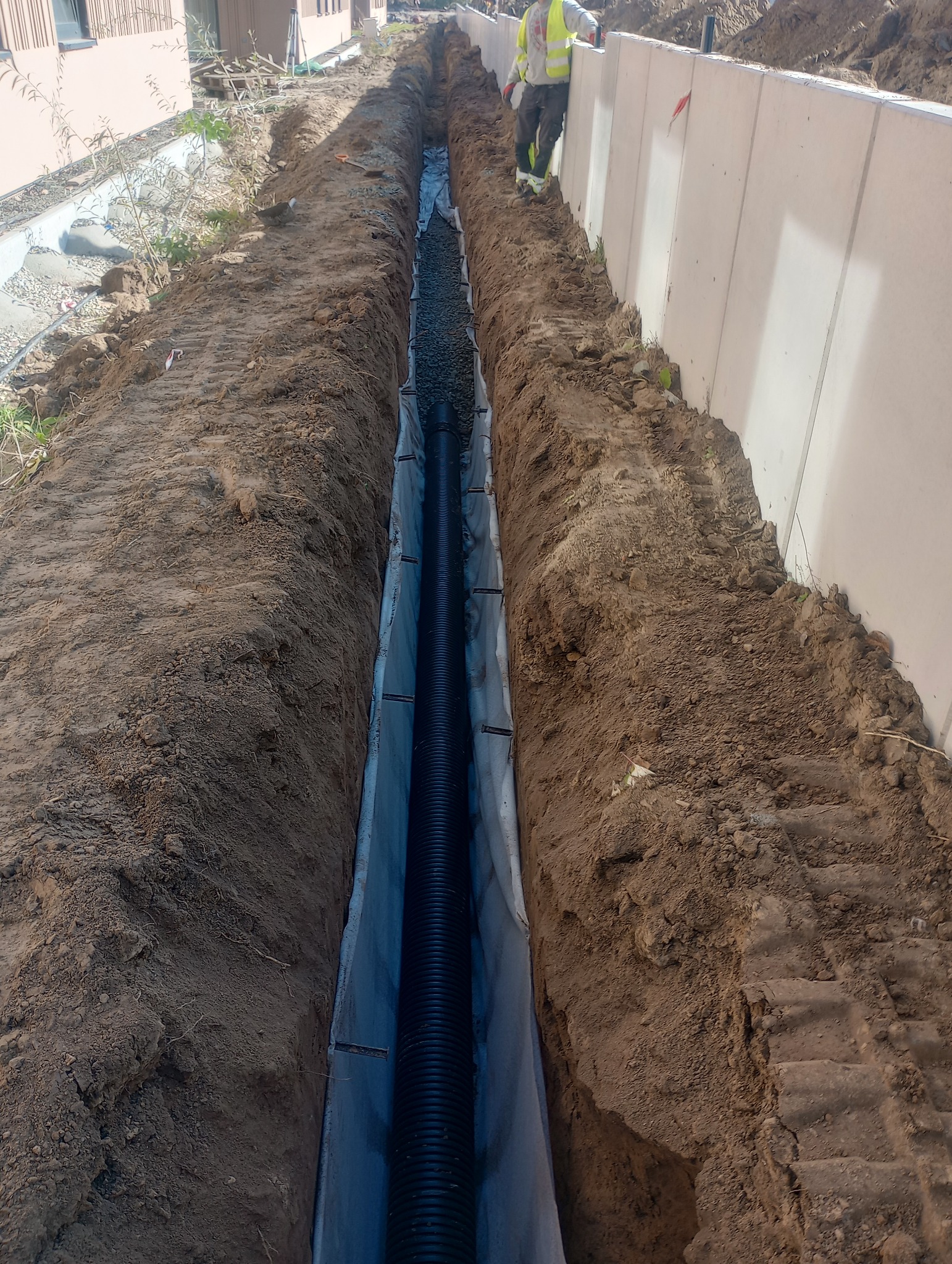 Długi, wąski wykop z czarną rurą drenażową ułożoną na geowłókninie i zasypaną kamieniami, widoczny fragment pracownika w odblaskowej kamizelce przy betonowym ogrodzeniu.