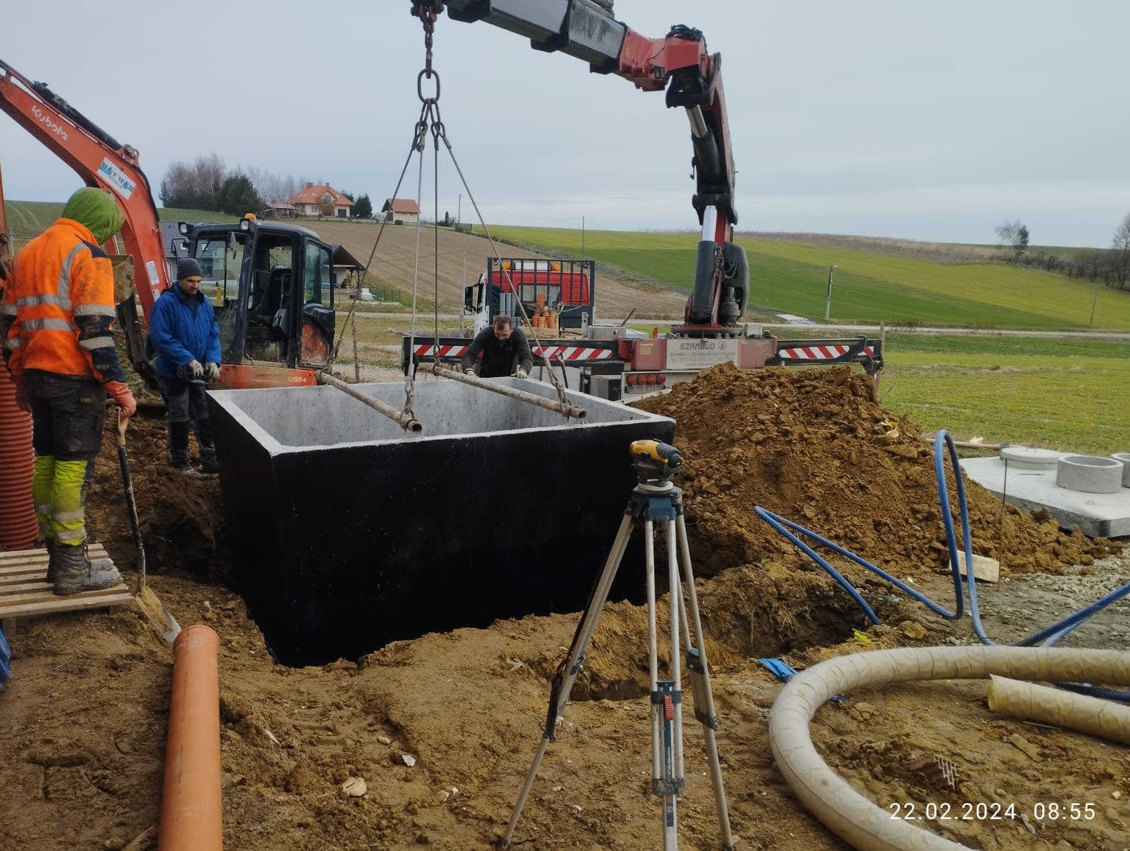 Montaż betonowego zbiornika z pomocą dźwigu: pracownicy w odblaskowych ubraniach nadzorują opuszczanie elementu do wykopu, w tle widoczny krajobraz wiejski i maszyny budowlane.