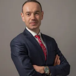 Portret mężczyzny w granatowym garniturze w kratę, białej koszuli i czerwonym krawacie z wzorem paisley, pozującego z założonymi rękami i zegarkiem na tle szarego studia.