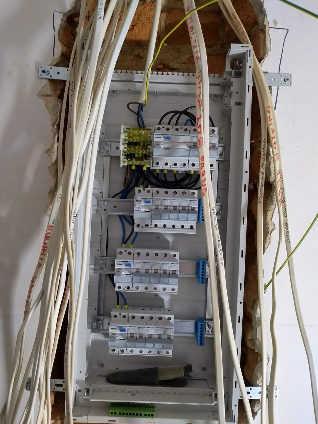 Rozdzielnica elektryczna w trakcie montażu, widoczne przewody opisane markerem, wystające ze ściany, obok fragment odsłoniętej cegły.