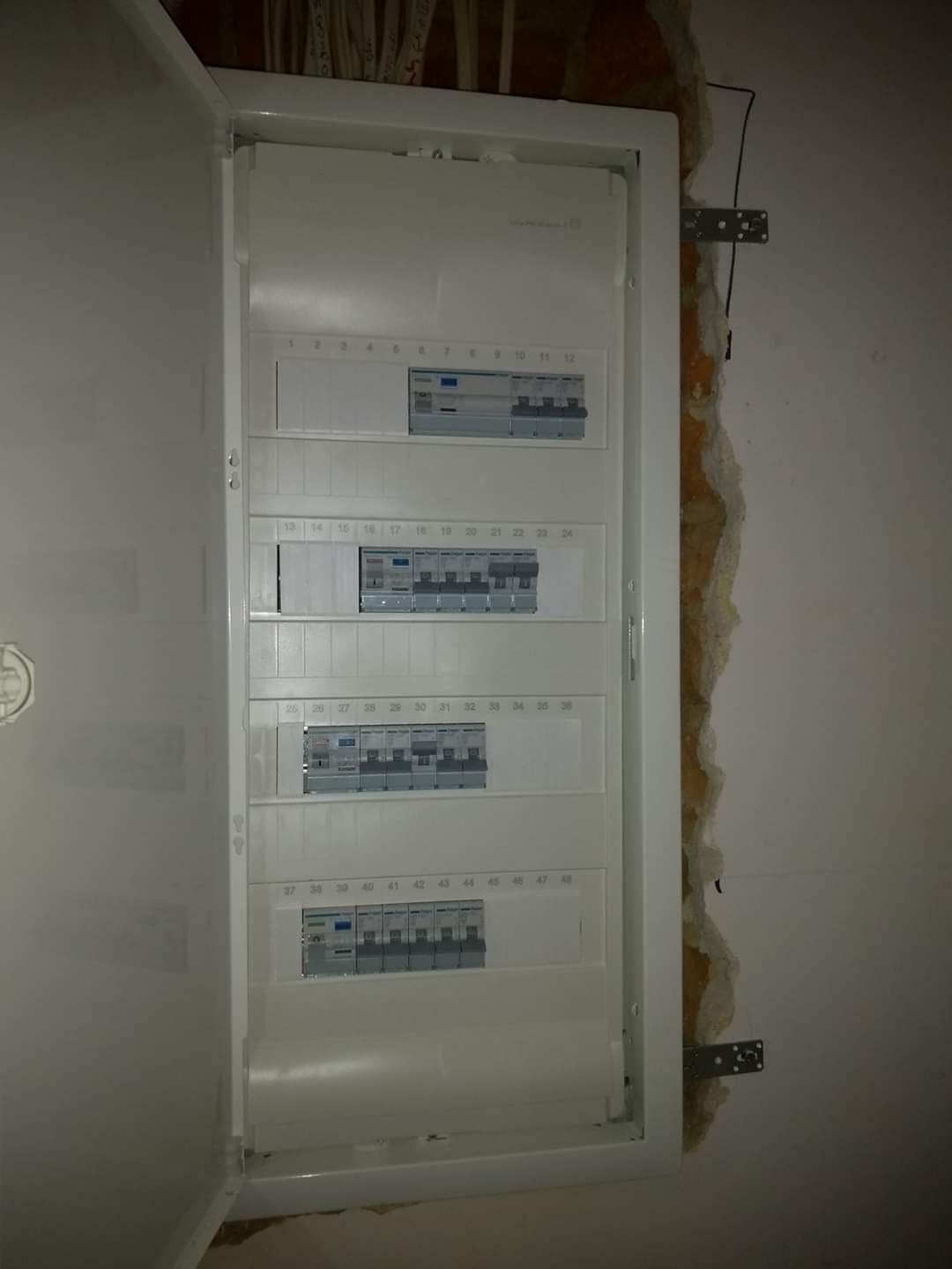 Otwarta skrzynka rozdzielcza z bezpiecznikami na ścianie w trakcie montażu, widoczne kable w suficie i surowa ściana obok.