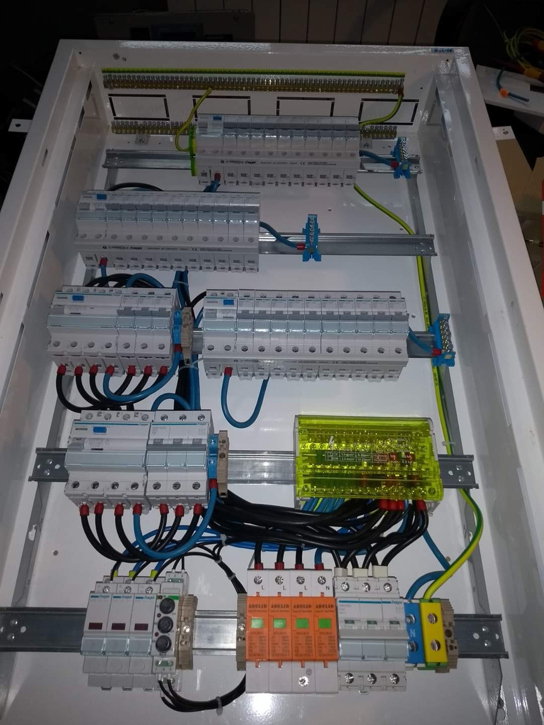 Wewnętrzny widok rozdzielnicy elektrycznej z zabezpieczeniami różnicowoprądowymi, ogranicznikami przepięć (pomarańczowe) i żółtym modułem, zamontowanej w białej obudowie.