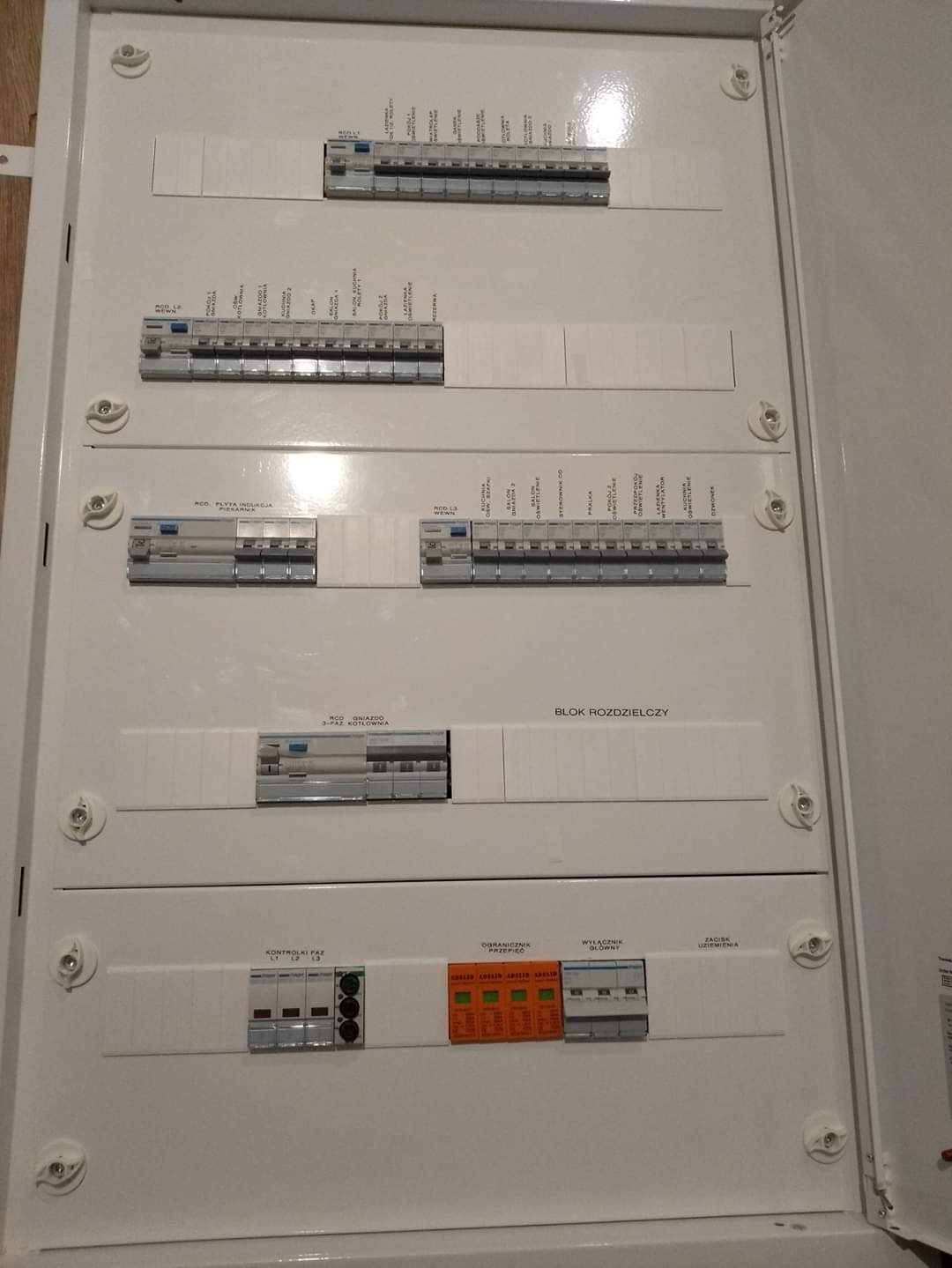 Otwarta, biała skrzynka rozdzielcza z bezpiecznikami i wyłącznikami, widoczne etykiety opisujące obwody, ujęcie z bliska, światło odbija się od powierzchni.