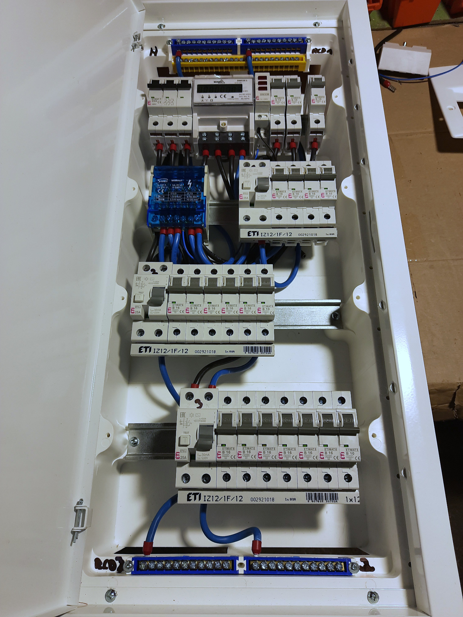 Otwarta skrzynka bezpiecznikowa z zainstalowanymi bezpiecznikami ETI IZ12/1F/12, widoczne przewody i listwy zaciskowe, całość w białej obudowie.