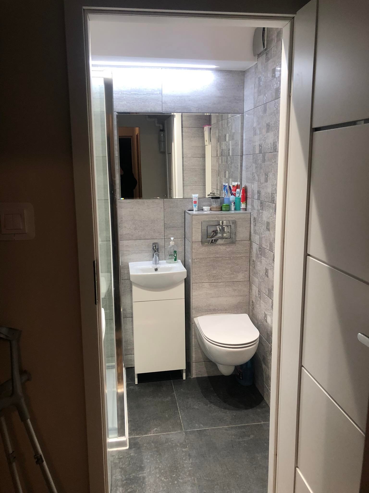 Ukończona łazienka z szarymi płytkami, wiszącą toaletą, białą umywalką z szafką i lustrem, widoczna zza otwartych drzwi; w tle fragment kabiny prysznicowej.