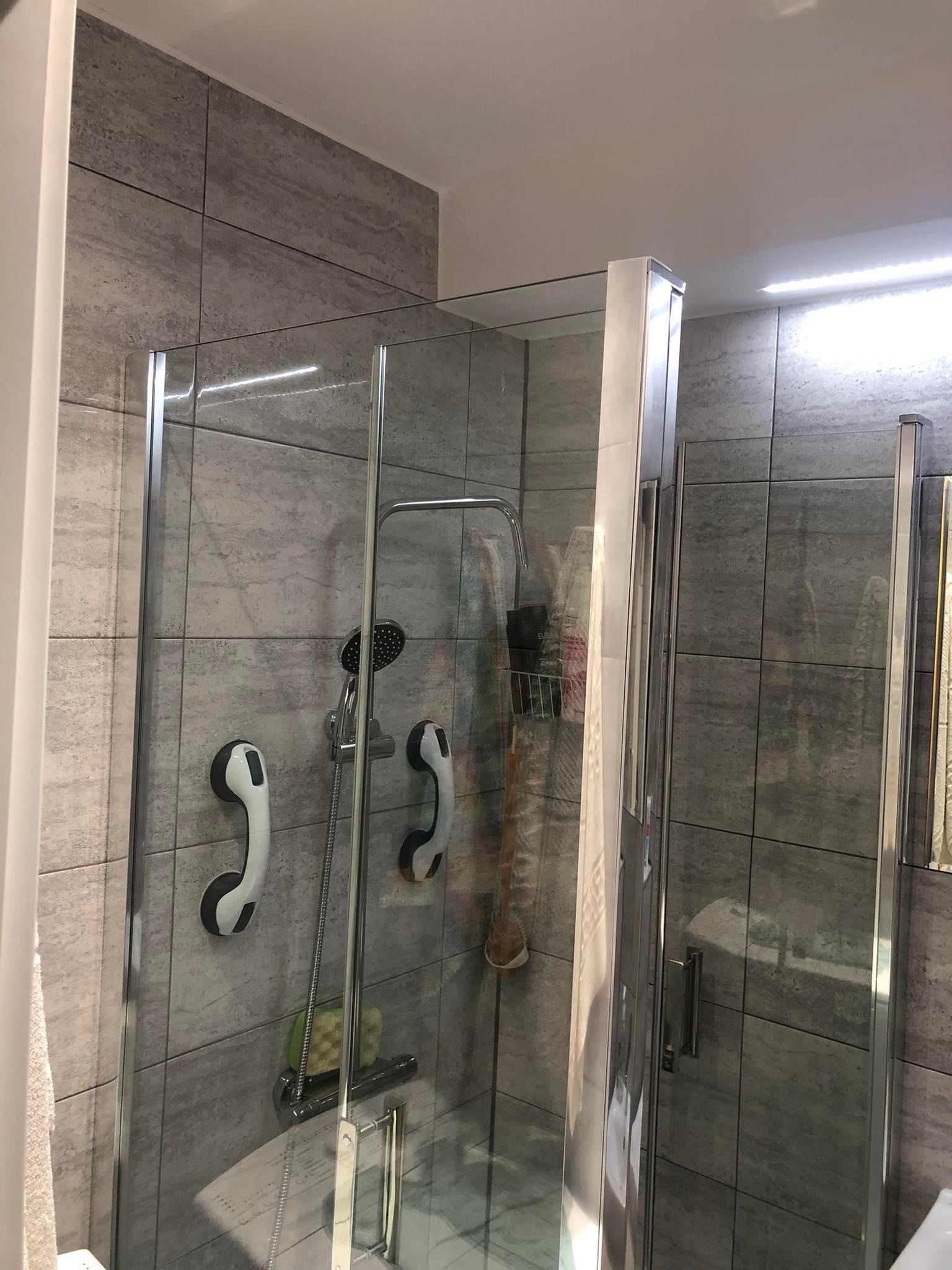 Szklana kabina prysznicowa z chromowanymi profilami, zamontowana w łazience wyłożonej szarymi płytkami imitującymi kamień, z zamontowanymi uchwytami dla osób z niepełnosprawnościami.