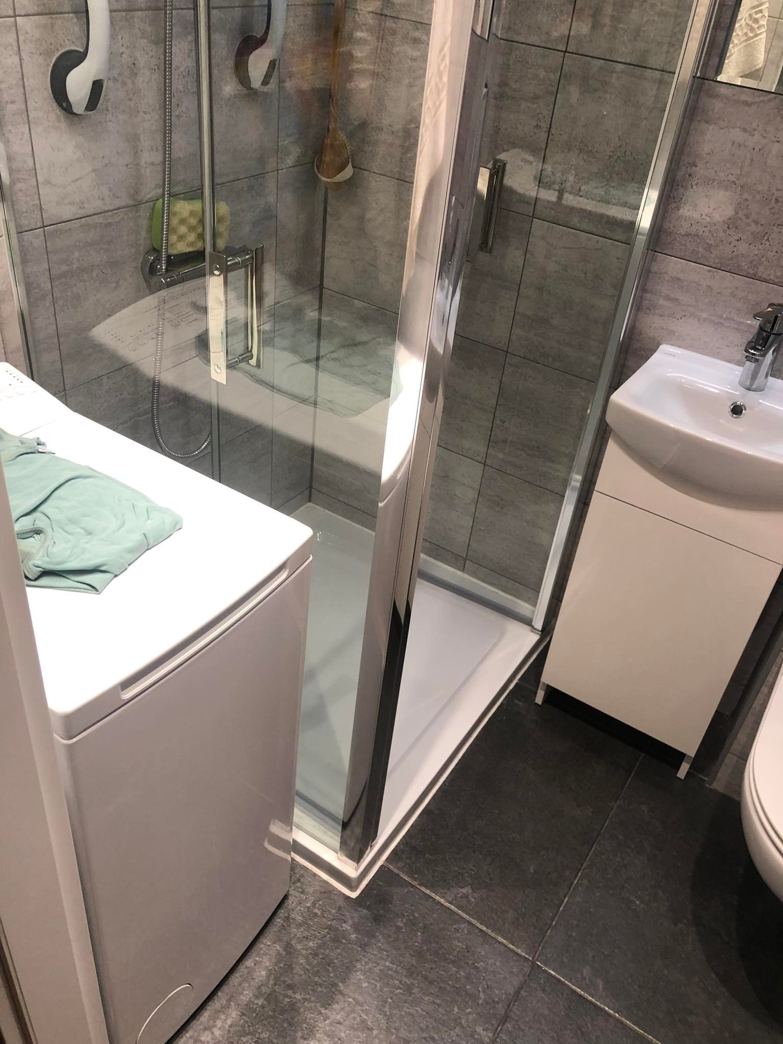 Nowoczesna łazienka z szarymi płytkami, kabiną prysznicową ze srebrnymi profilami, białą umywalką z szafką, pralką z turkusowym ręcznikiem i ciemną podłogą.