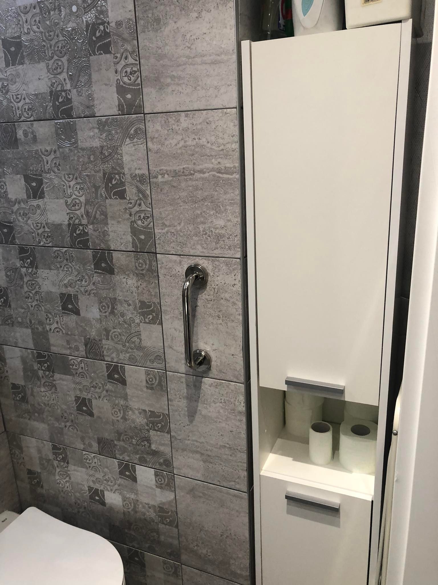 Uchwyt bezpieczeństwa zamontowany na ścianie z szarych płytek o zróżnicowanych wzorach, obok biała szafka łazienkowa z zapasem papieru toaletowego.