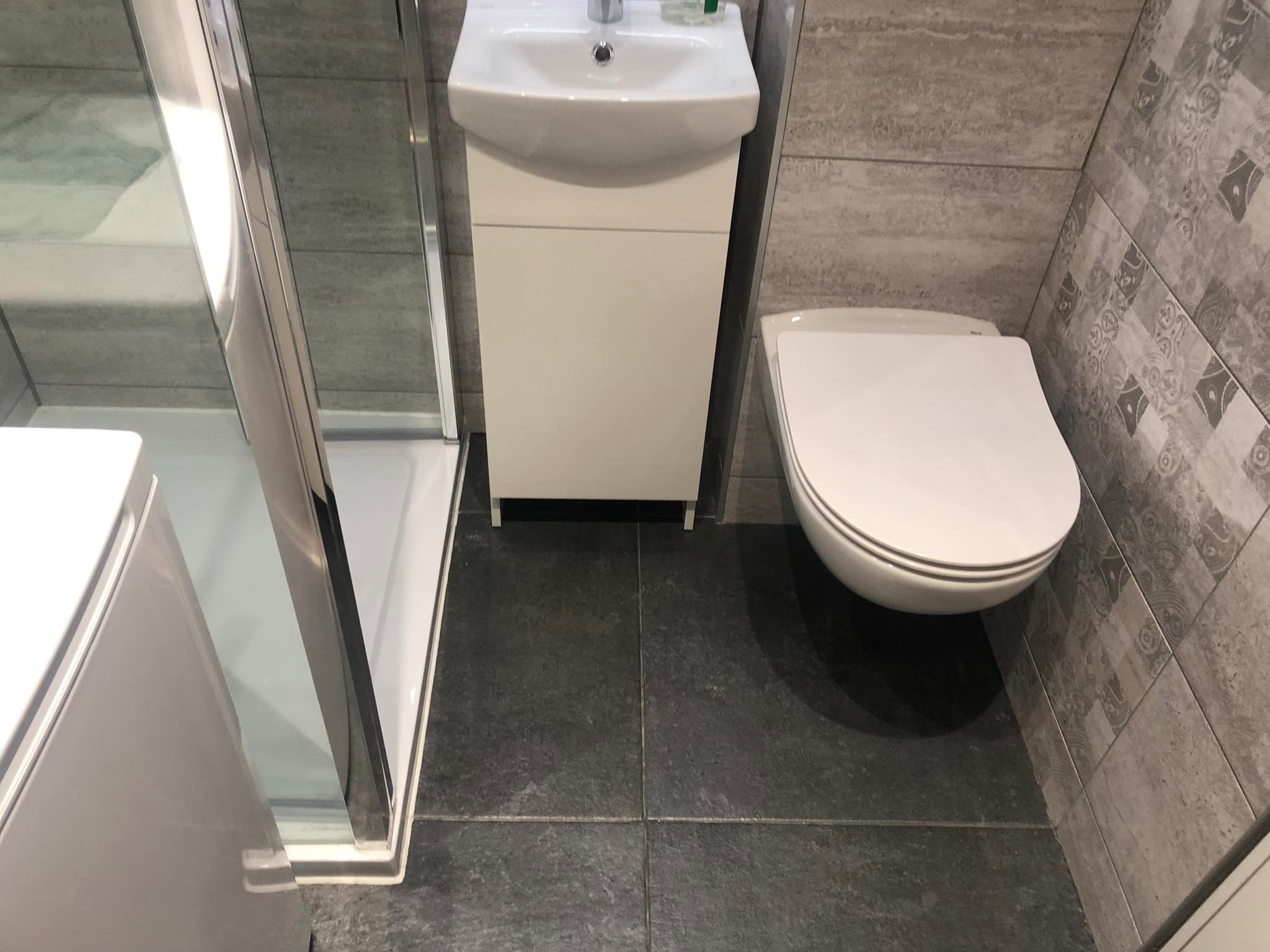 Nowoczesna łazienka z kabiną prysznicową, podwieszaną toaletą i umywalką z szafką, wykończona szarymi płytkami o zróżnicowanym wzorze i ciemną podłogą.