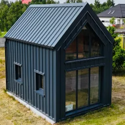 Nowoczesny, ciemnoszary domek z blachy na fundamencie, z dużymi oknami i dwuspadowym dachem, otoczony zielenią. Widok z zewnątrz.