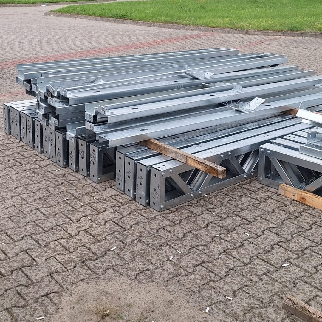 Stalowe, ocynkowane elementy konstrukcyjne domu szkieletowego ułożone na betonowej kostce brukowej, widoczne otwory montażowe i wzmocnienia poprzeczne.