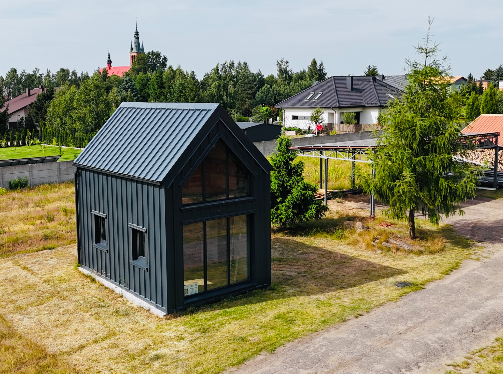Czarny, nowoczesny dom modułowy z blachy na tle zieleni i zabudowań. Widoczne okna, blaszany dach i elewacja. W tle kościół z czerwoną wieżą.