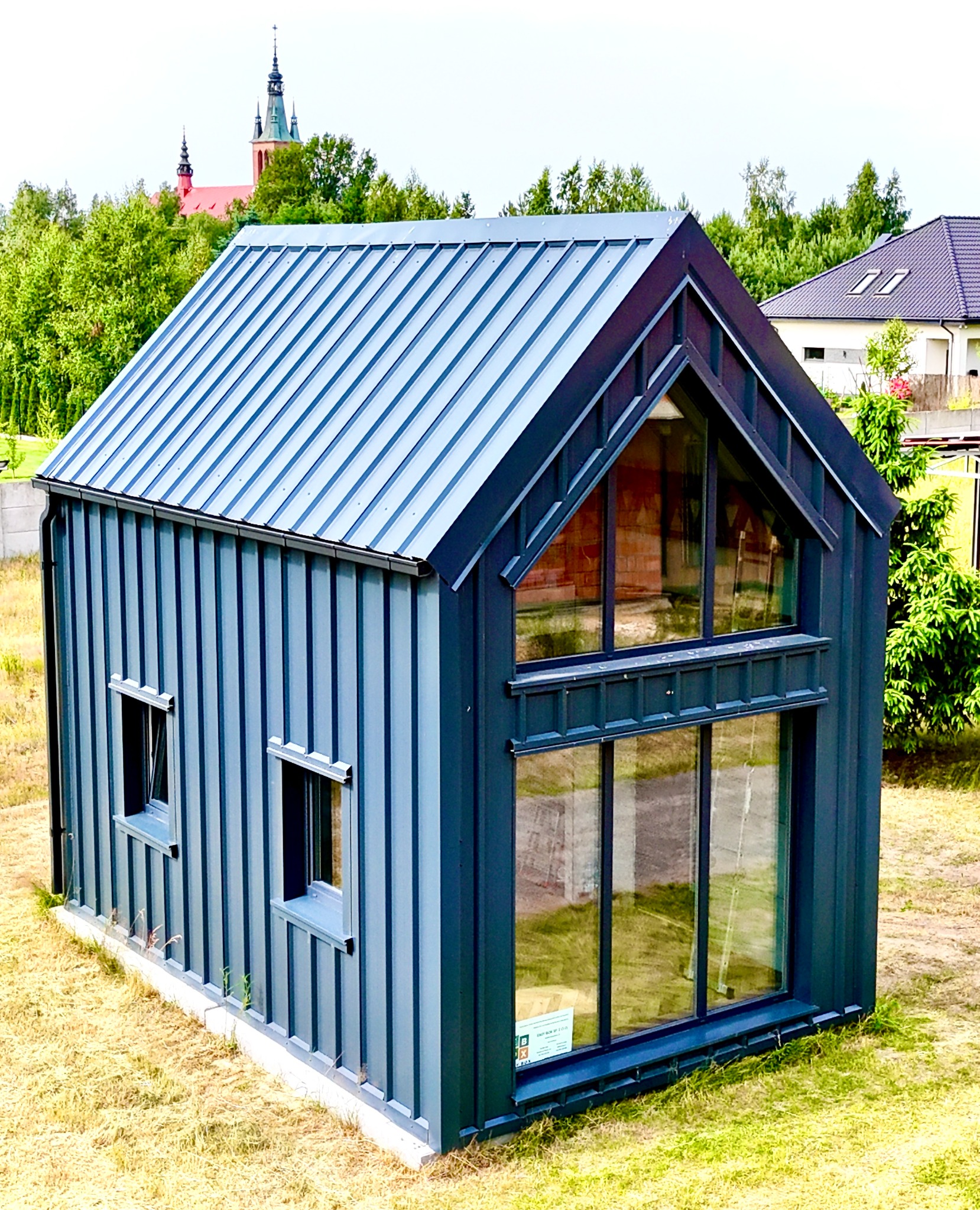 Niebieski, nowoczesny dom z blachy trapezowej z dużymi oknami i dwuspadowym dachem na tle zieleni i kościoła. Widoczne detale wykończenia i elementy architektoniczne.