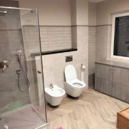 Nowoczesna łazienka z kabiną prysznicową typu walk-in, wiszącą toaletą i bidetem, wykończona płytkami w odcieniach szarości i beżu z drewnopodobną podłogą ułożoną w jodełkę.