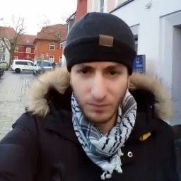 Selfie mężczyzny w czapce i szaliku keffiyeh na tle miejskiego krajobrazu z budynkami i zaparkowanymi samochodami.