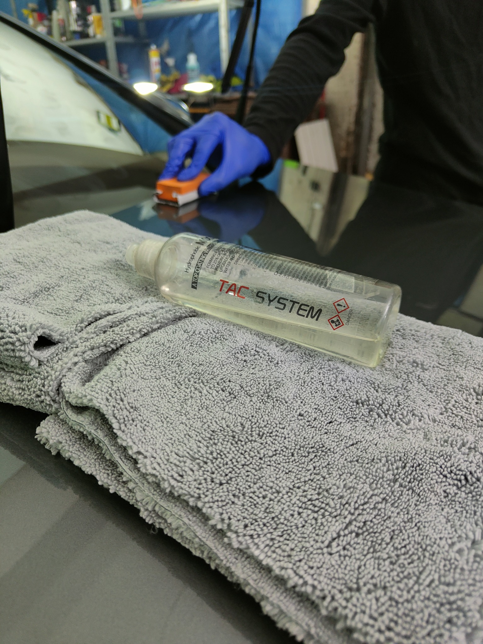 Nakładanie powłoki ochronnej na lakier samochodu za pomocą aplikatora i preparatu TAC System, obok leży złożona szara mikrofibra.
