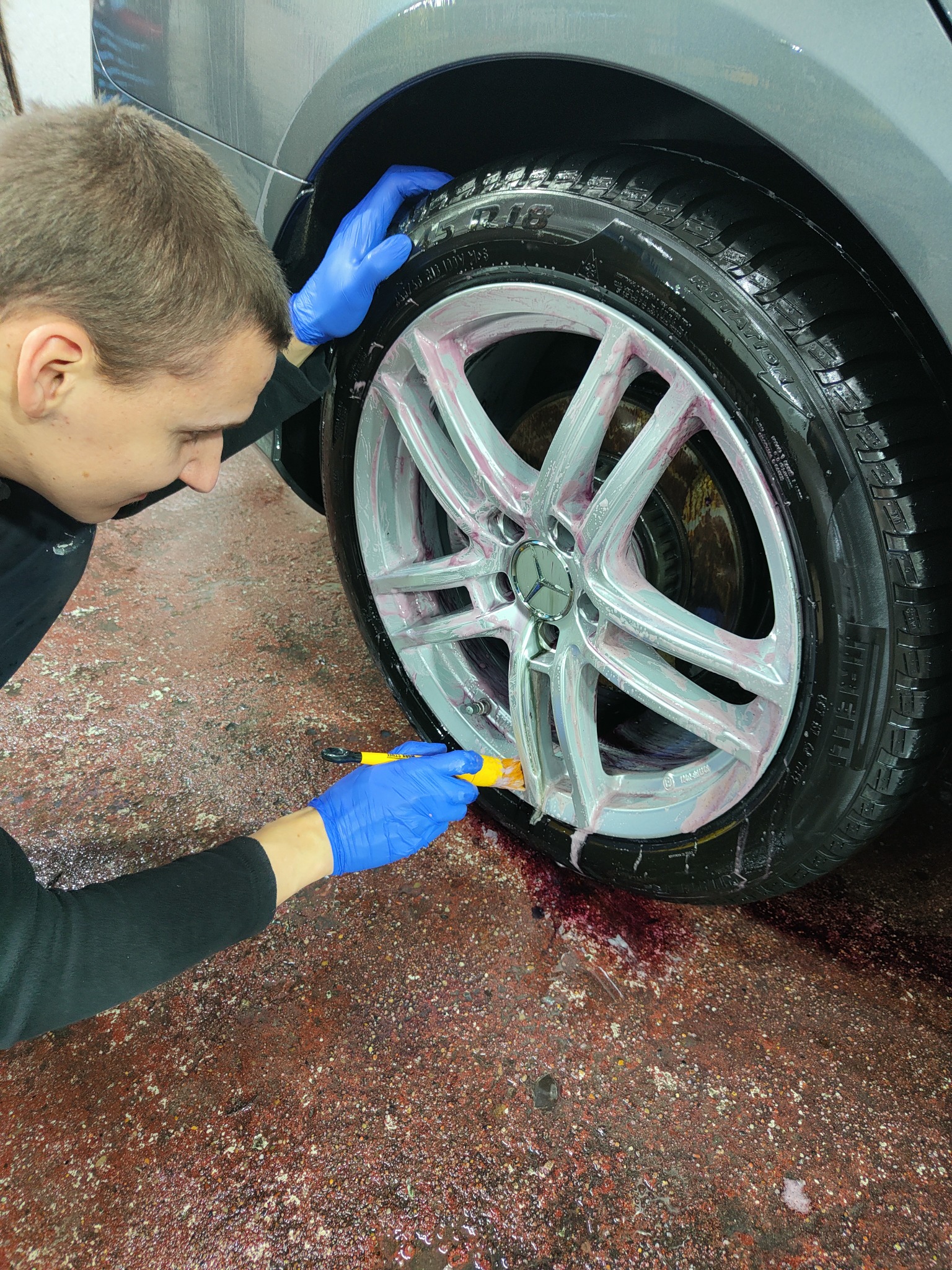 Czyszczenie aluminiowej felgi Mercedes-Benz szczoteczką z żółtą rączką, widoczny czerwony płyn spływający po feldze i oponie na brudną posadzkę myjni.