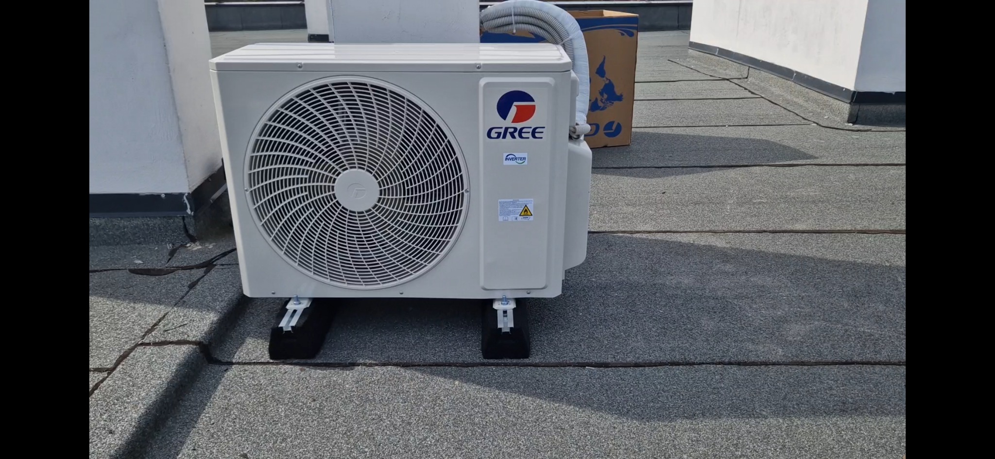 Biała jednostka zewnętrzna klimatyzacji marki Gree, zamontowana na czarnych podkładkach na płaskim dachu pokrytym papą.
