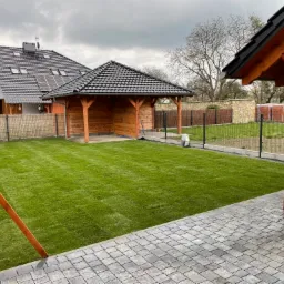 Świeżo założony trawnik z rolki w ogrodzie, widoczna drewniana altana z czarnym dachem, kostka brukowa i dom z panelami słonecznymi w tle.
