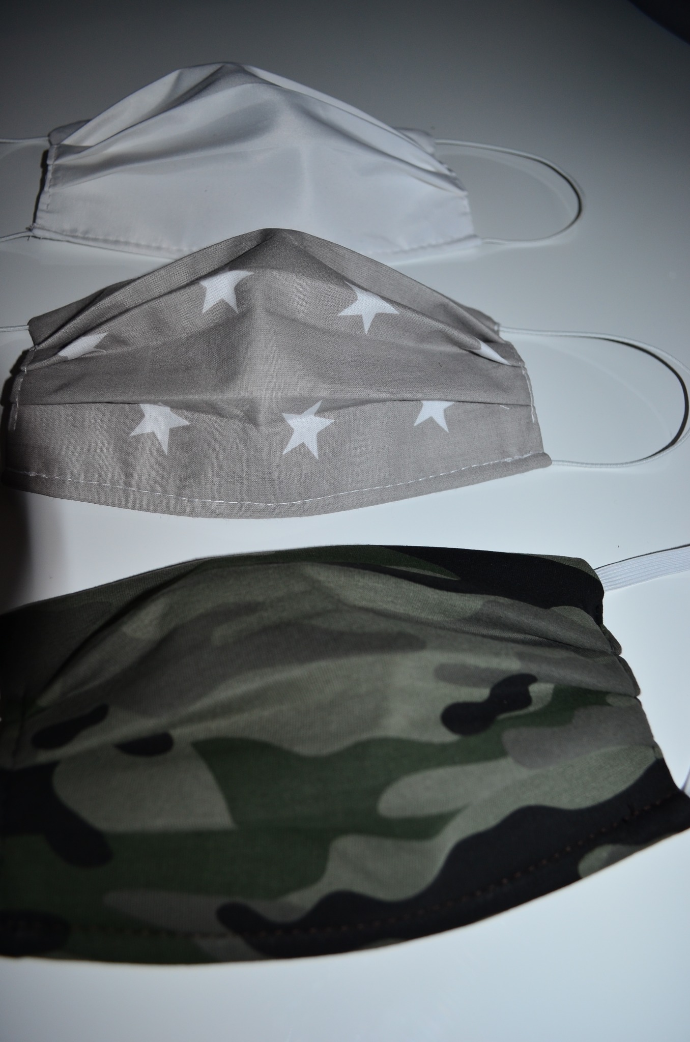 Trzy maseczki ochronne leżące jedna na drugiej: biała, szara w białe gwiazdki i moro, wszystkie z gumkami.