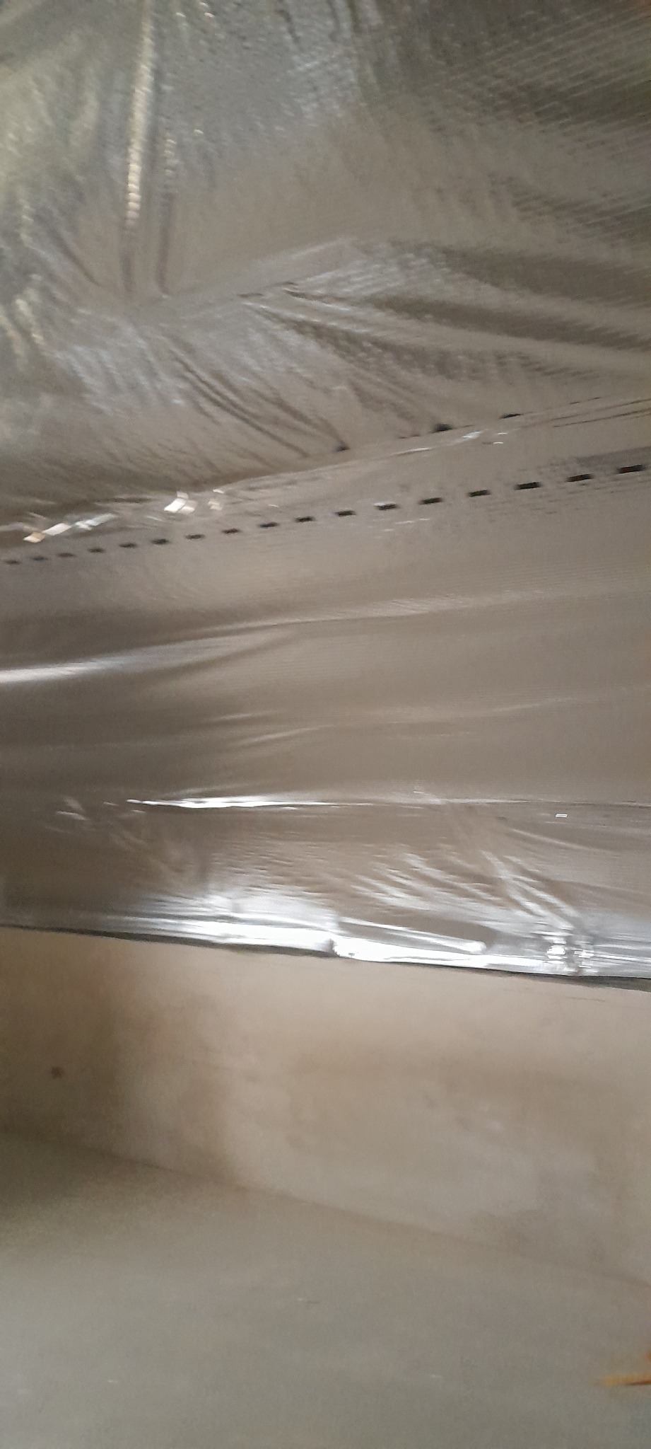 Błyszcząca folia paroizolacyjna, przymocowana zszywkami do drewnianych łat na suficie, widoczna surowa ściana poniżej.