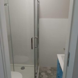 Emilian Fedorowicz Usługi remontowo budowlane - Wyremontowana łazienka z kabiną prysznicową z przesuwnymi drzwiami, toaletą, umywalką w niebieskiej szafce i podłogą wyłożoną szarymi heksagonalnymi płytkami.