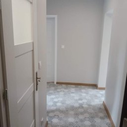 Emilian Fedorowicz Usługi remontowo budowlane - Korytarz po remoncie z szarymi ścianami, heksagonalną płytką podłogową w odcieniach szarości i bieli, oraz białymi drzwiami z przeszkleniem.