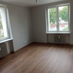 Emilian Fedorowicz Usługi remontowo budowlane - Pomalowane na szaro puste pomieszczenie z panelami podłogowymi w odcieniach brązu, dwa okna z białymi ramami i grzejnikami pod nimi, goła żarówka zwisająca z sufitu.