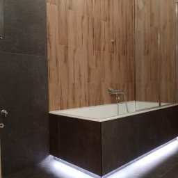 Emilian Fedorowicz Usługi remontowo budowlane - Wykończona łazienka z wanną obudowaną ciemnymi płytami imitującymi kamień, podświetloną listwą LED, drewnopodobnymi płytkami na ścianie i szklanym parawanem nawannowym.